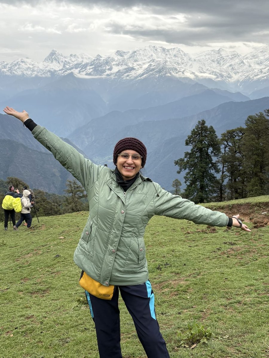 Harpreet Kaur Arora standing on a hillside with her arms outstretched, snow-covered Himalayan peaks behind her