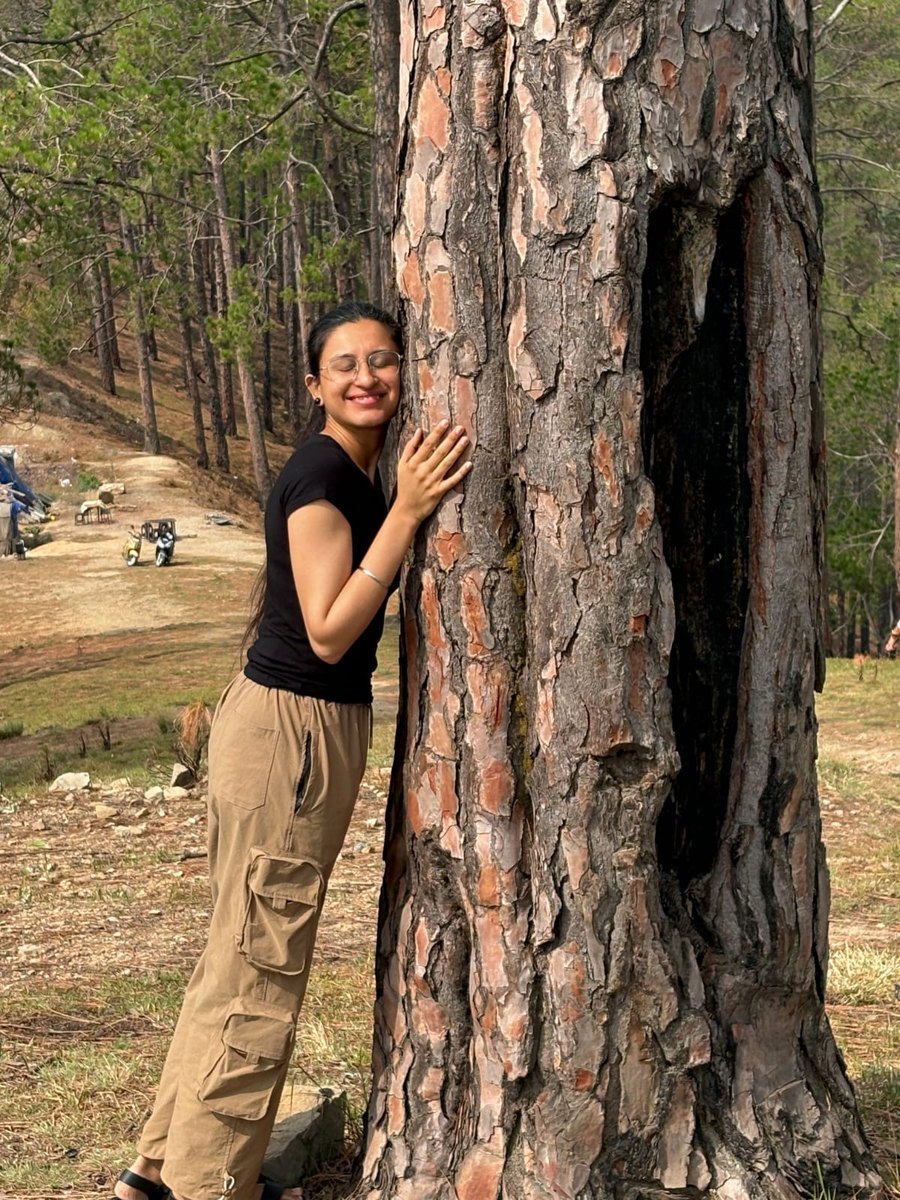 Harpreet Kaur Arora resting against the trunk of a pine tree in a hillside forest, eyes closed and smiling