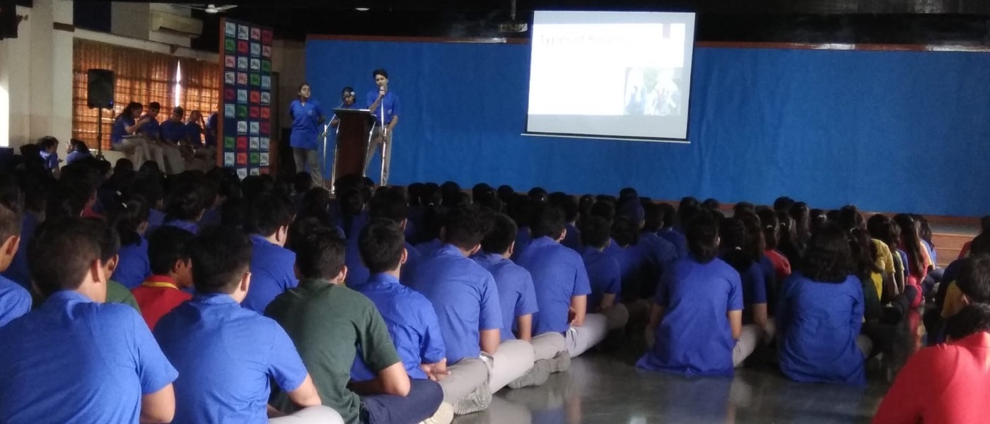 A school assembly hall filled with students seated on the floor, watching a workshop presentation on types of bullying